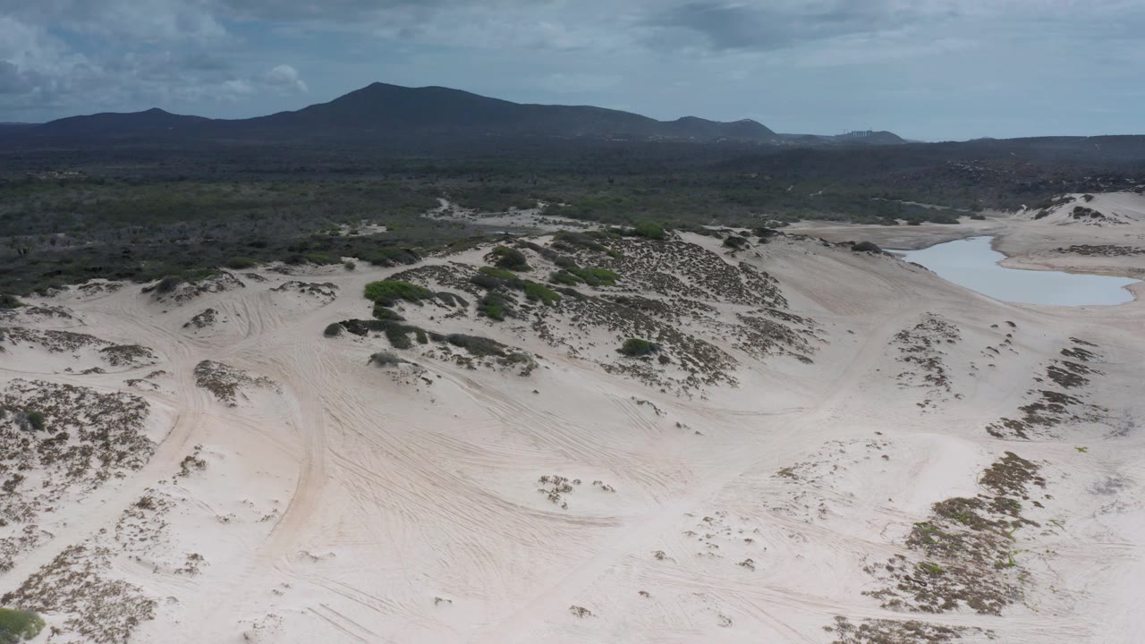 Aerial frame in Los Cabos, Mexico, with sandy hills crossing the foreground, green scrub patches, the pond set farther right, distant mountains under a clouded sky, and no visible people.