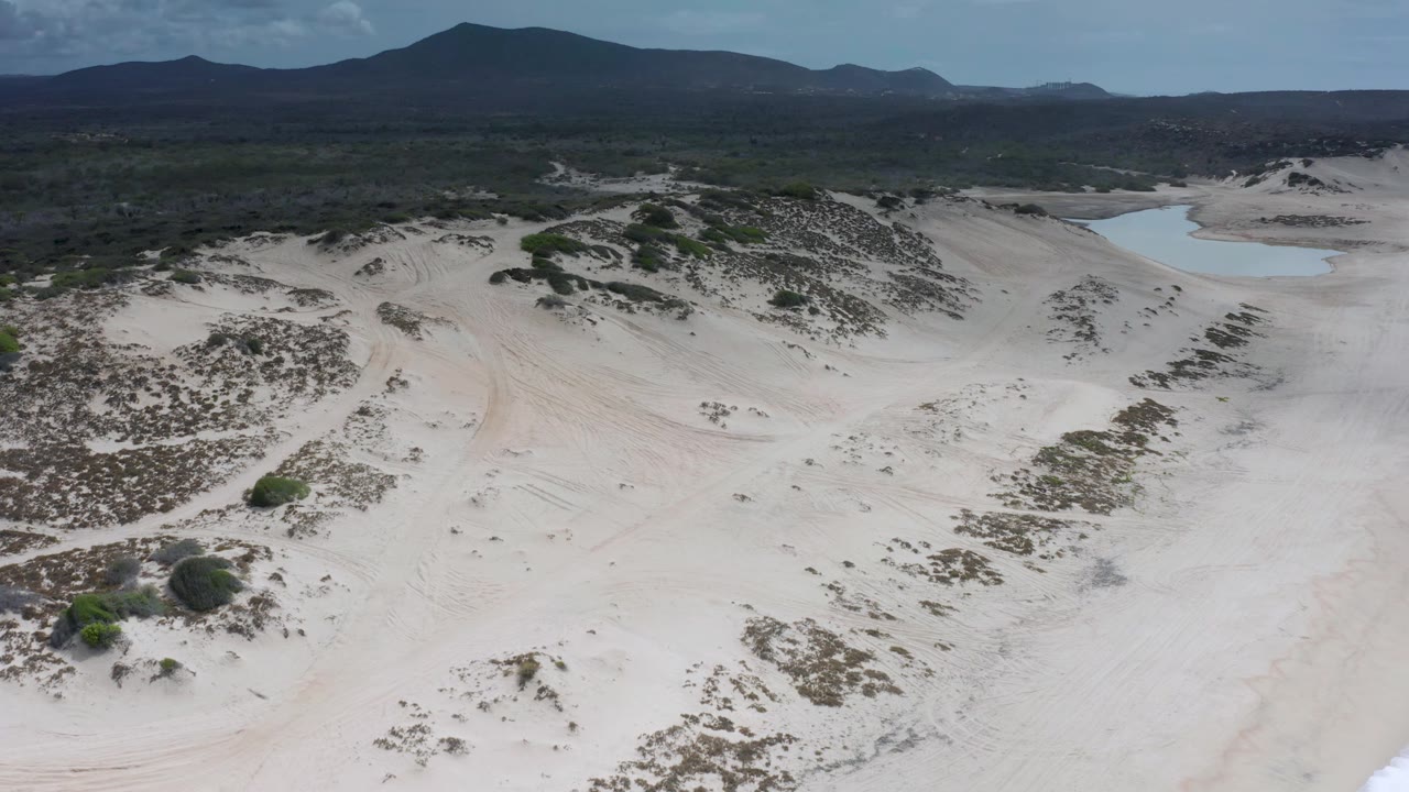 High aerial frame over Los Cabos, Mexico sand dunes with scattered scrub, a small pale blue inland pool near the upper right, distant hills, and no visible people.