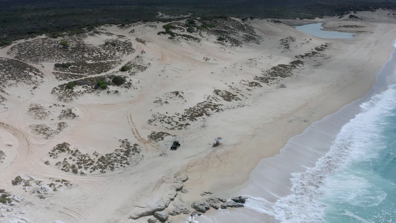 Drone frame over Los Cabos sand dunes with sparse vegetation, vehicle tracks, a rocky lower shoreline, blue-green water to the right, and no visible people.