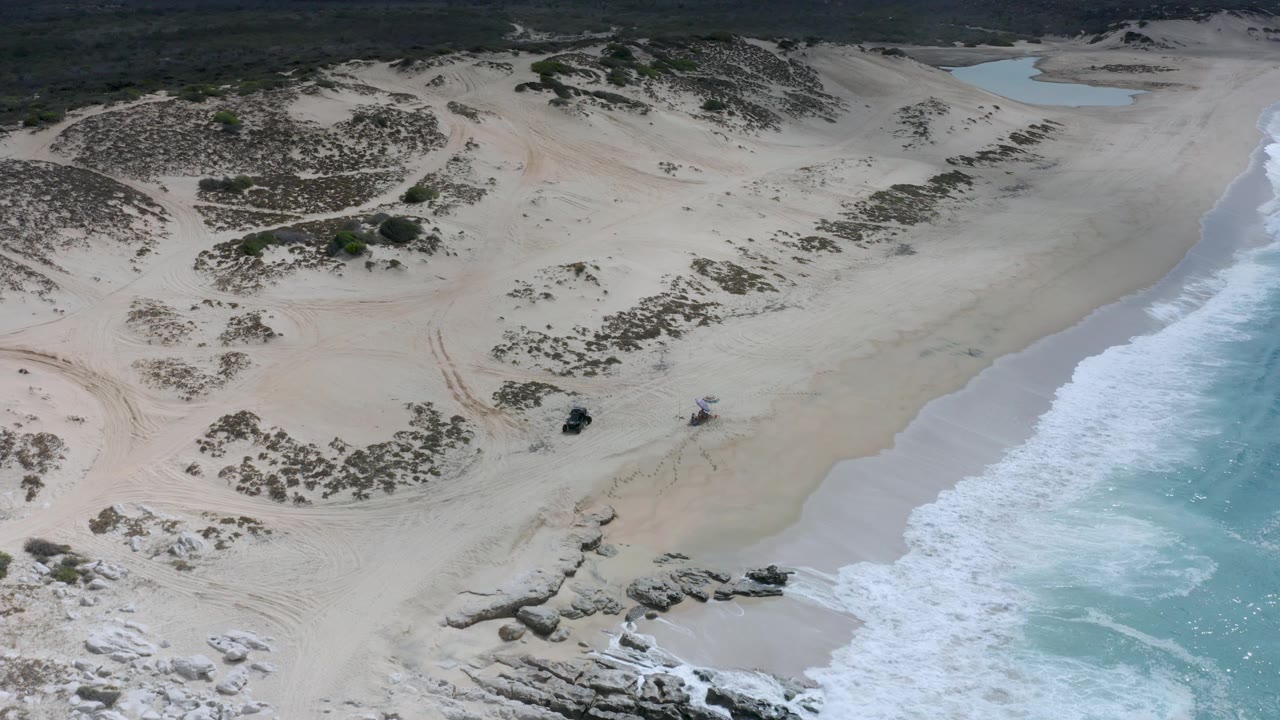 Aerial view of the Los Cabos coastline with a wider band of beach, scrub-covered dunes, rocky outcrops near the water, bright surf foam, and no visible people.