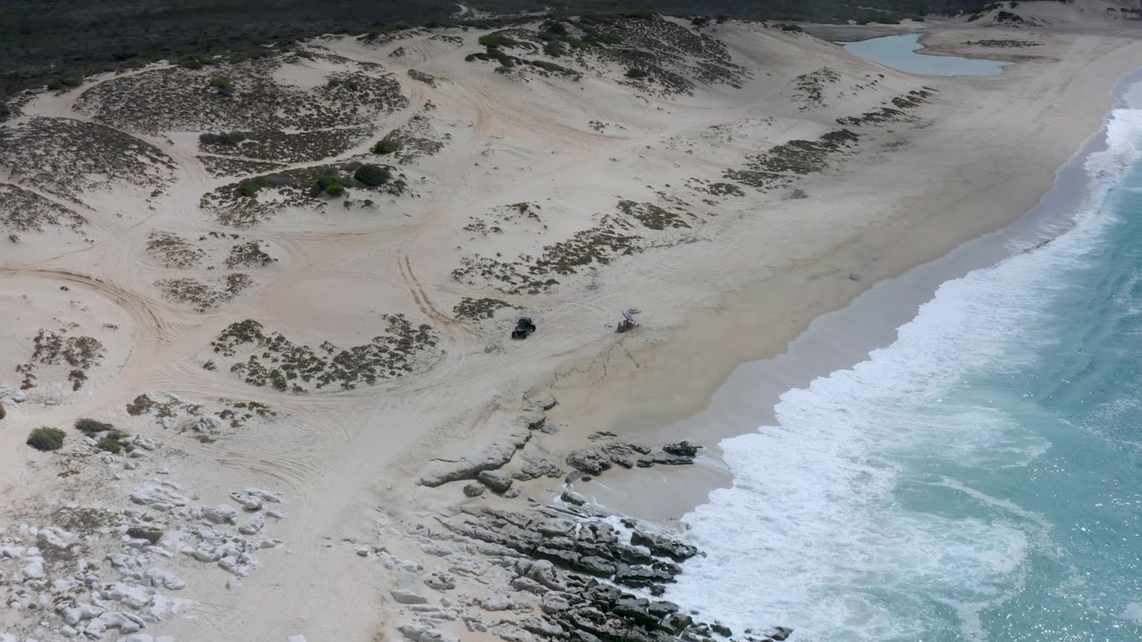Overhead Los Cabos beach frame with scrubby dunes across the upper half, a rocky point near the bottom, breaking waves at right, and no visible people.