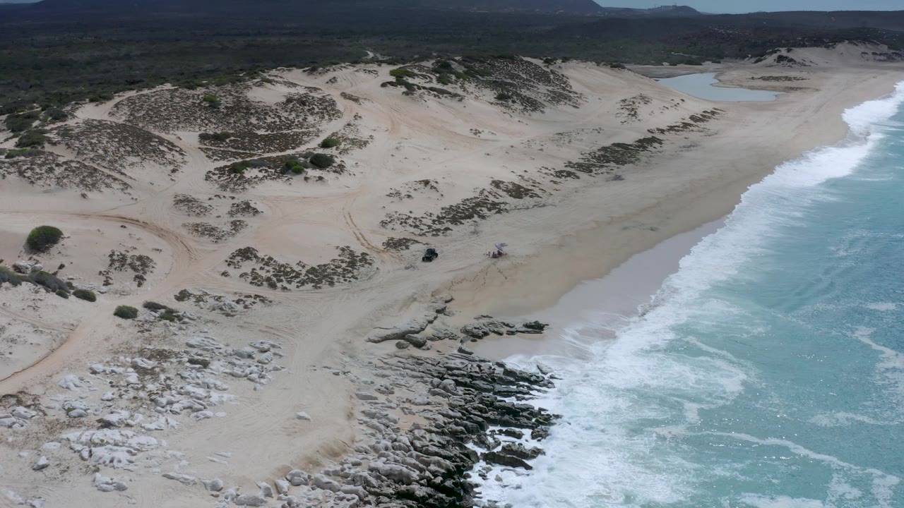 Drone-like view above the Los Cabos coast with turquoise water and foam on the right, dark shoreline rocks below center, pale dunes inland, and no visible people.