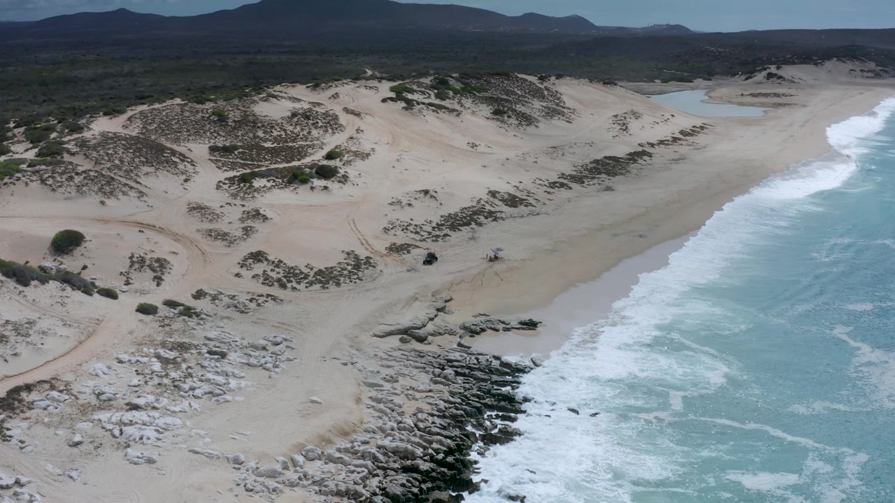 Wide aerial view of a Los Cabos beach with the surf band on the right, dune ridges across the center, a small inland water area near the horizon, and no visible people.