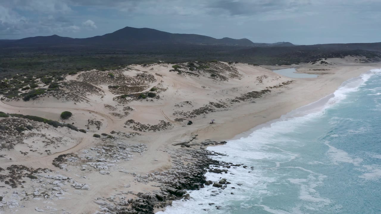 Wide aerial view in Los Cabos with the surf line running up the right edge, dune ridges across the center, inland scrub behind them, distant hills, and no visible people.