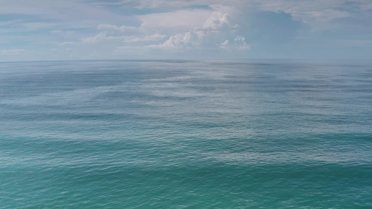 Blue green ocean surface stretches toward a soft horizon in Los Cabos, Mexico, with thin cloud cover and no visible people.