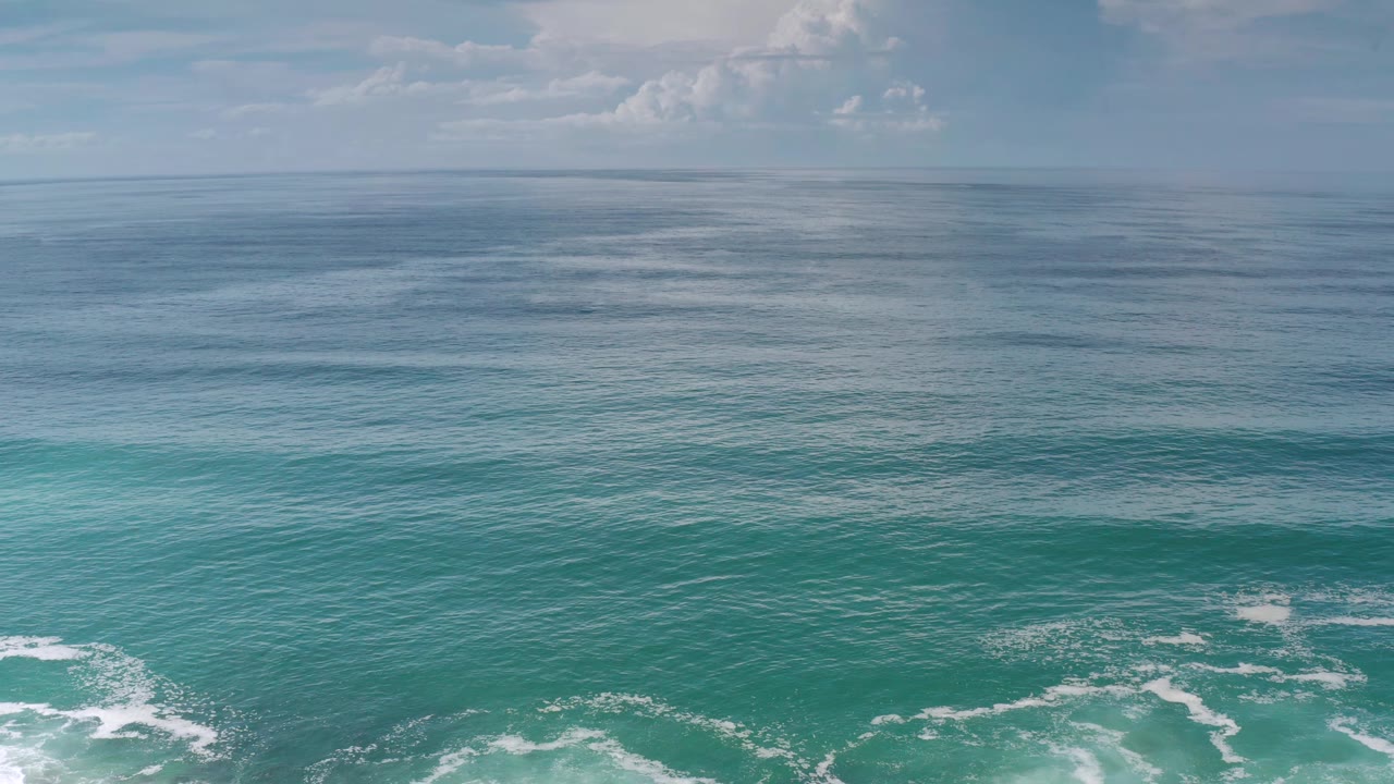 Los Cabos water fills the frame with darker blue water near the horizon, turquoise surf bands below, light cloud cover, and no visible people.