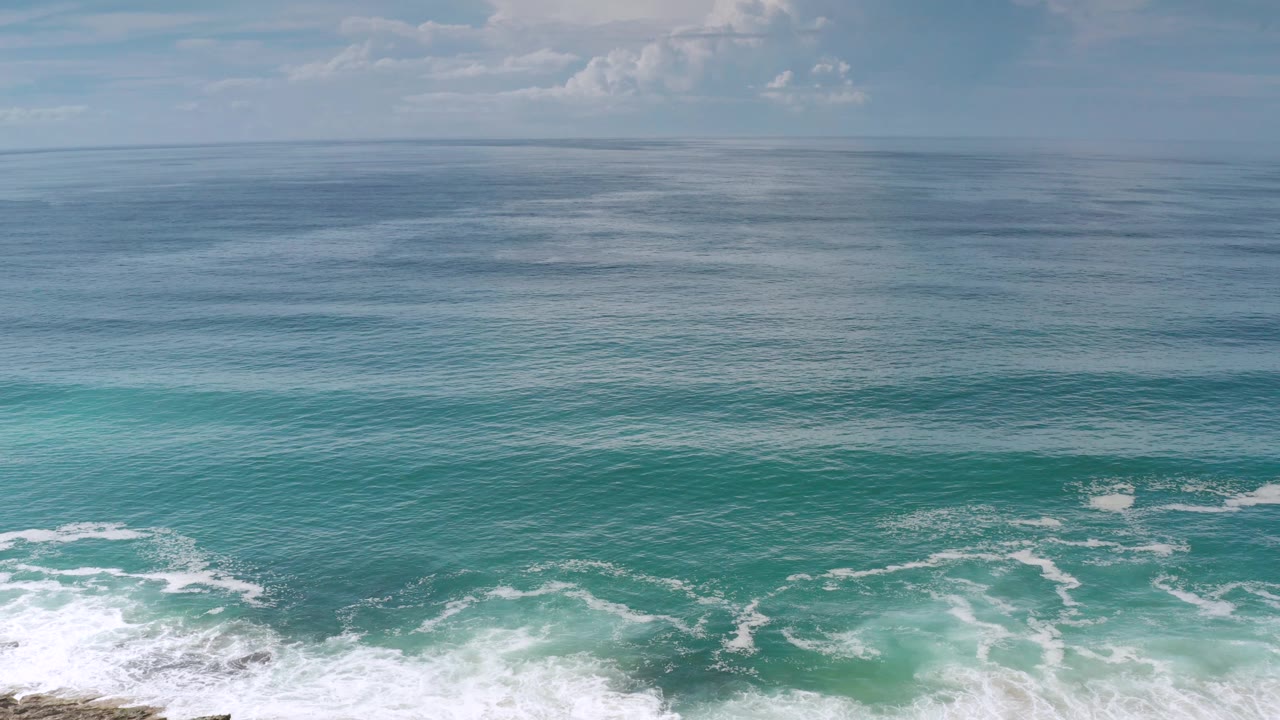 Los Cabos surf moves across blue-green water toward a pale sandy beach entering the lower frame, with open ocean and no visible people.