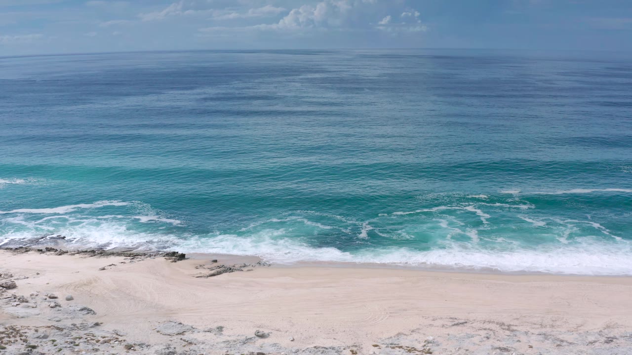 A second aerial frame in Los Cabos holds on rippling blue-green water, a bright surf band, pale beach, small shoreline rocks, and no visible people.