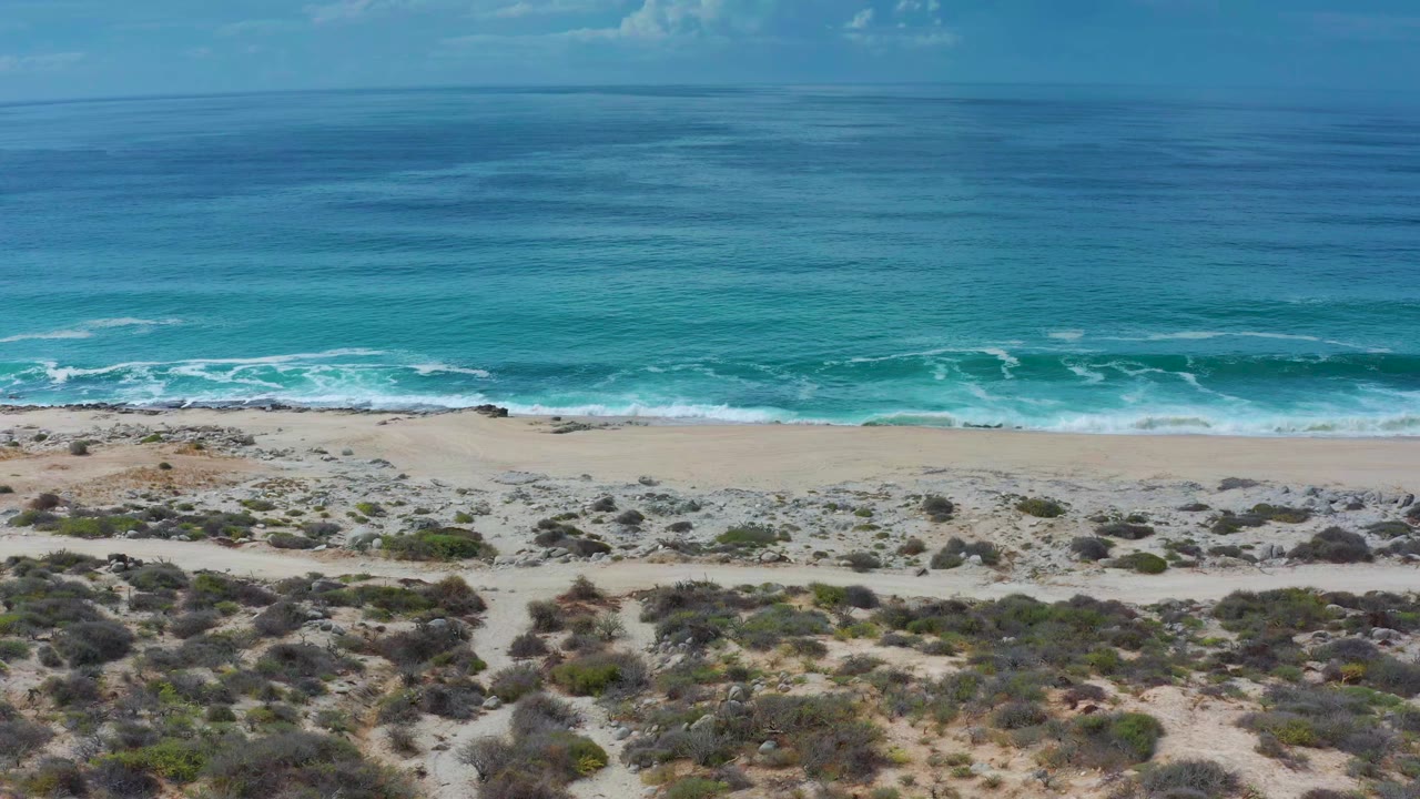 At 0:00, an elevated Los Cabos coastline view shows turquoise water, white surf, a pale beach, rocky sand, and low scrub with no visible people.