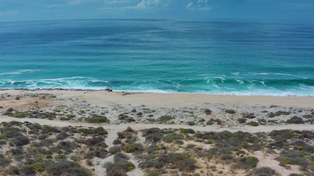 At 0:02, blue-green water fills the background while white foam marks the shore and dry scrubland covers the foreground, with no visible people.