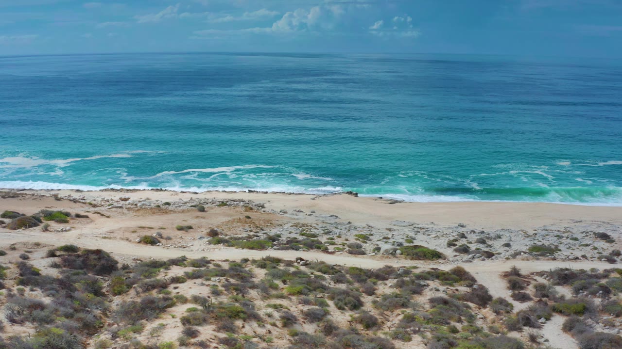 Los Cabos, Mexico shoreline seen from above with a wider band of turquoise water rolling toward the sand, low dune vegetation below, and no visible people.