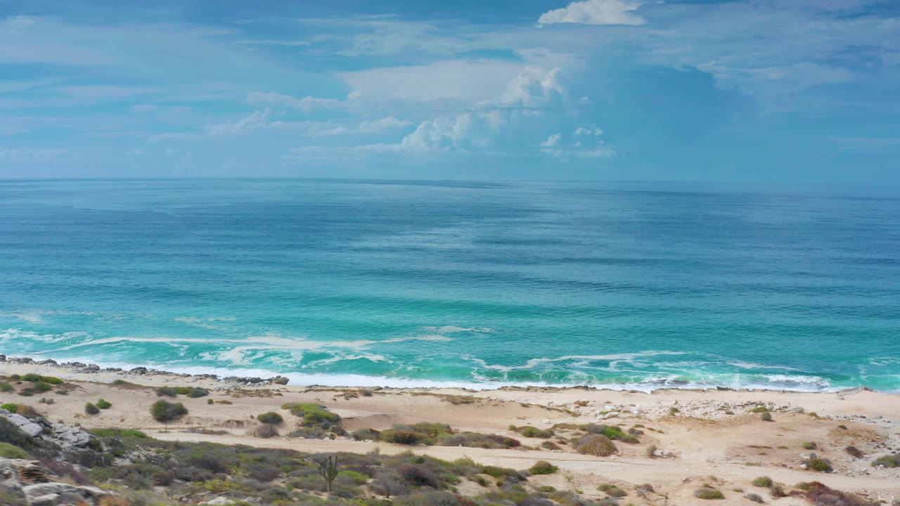 Wide ocean view with surf, sandy dunes, and clouds on the horizon.