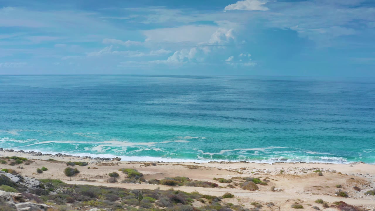 The Los Cabos shoreline remains in a wide frame as white breakers run along the beach below blue-green water and a clouded horizon, with no visible people.