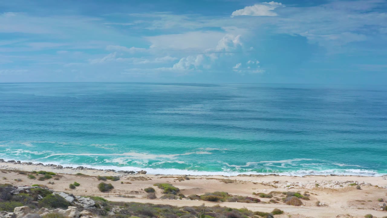 A wide Los Cabos coastal frame shows the surf band closer to the lower edge, open ocean across the center, distant clouds, and no visible people.