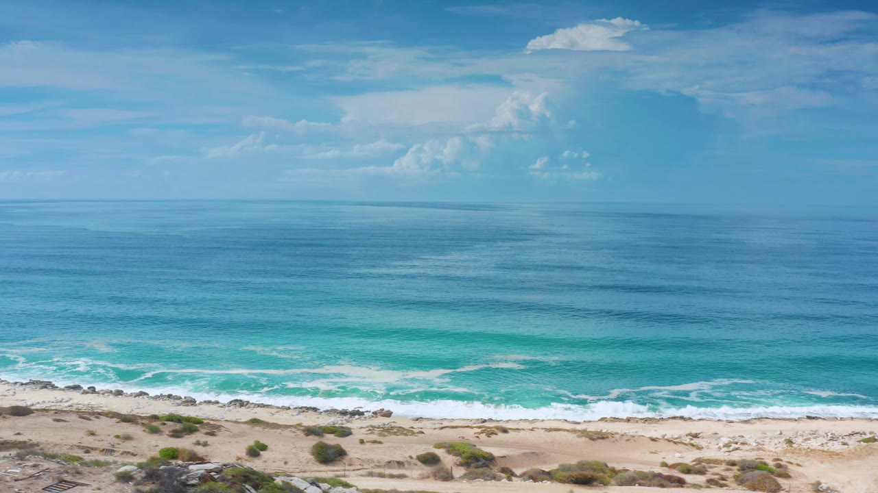 The Los Cabos coast appears empty as turquoise water rolls toward a sandy, rocky beach with dunes below and a tall cloud formation near the horizon.