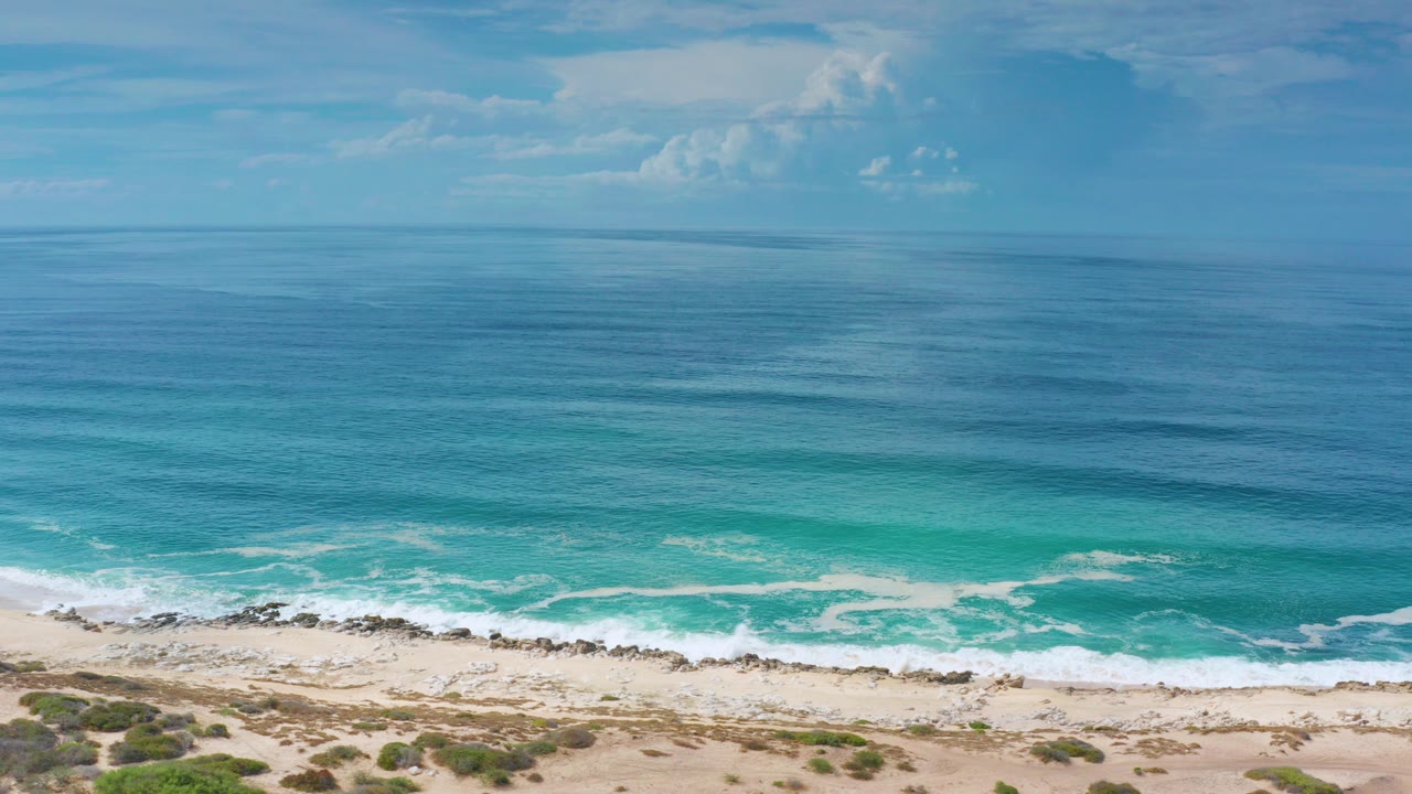 Aerial view of blue ocean, white surf, sandy beach, rocks, and coastal scrub.