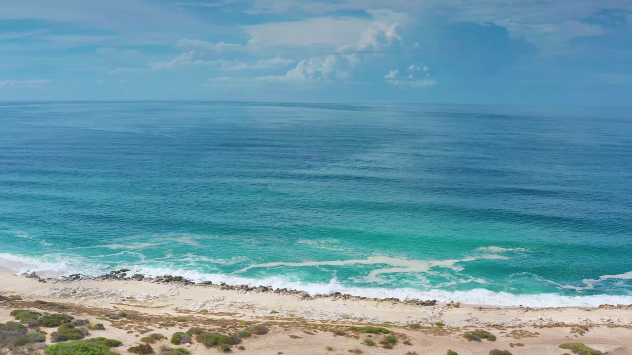The Los Cabos shoreline remains empty as white wave foam spreads across turquoise water above pale sand and low vegetation, with no visible people.