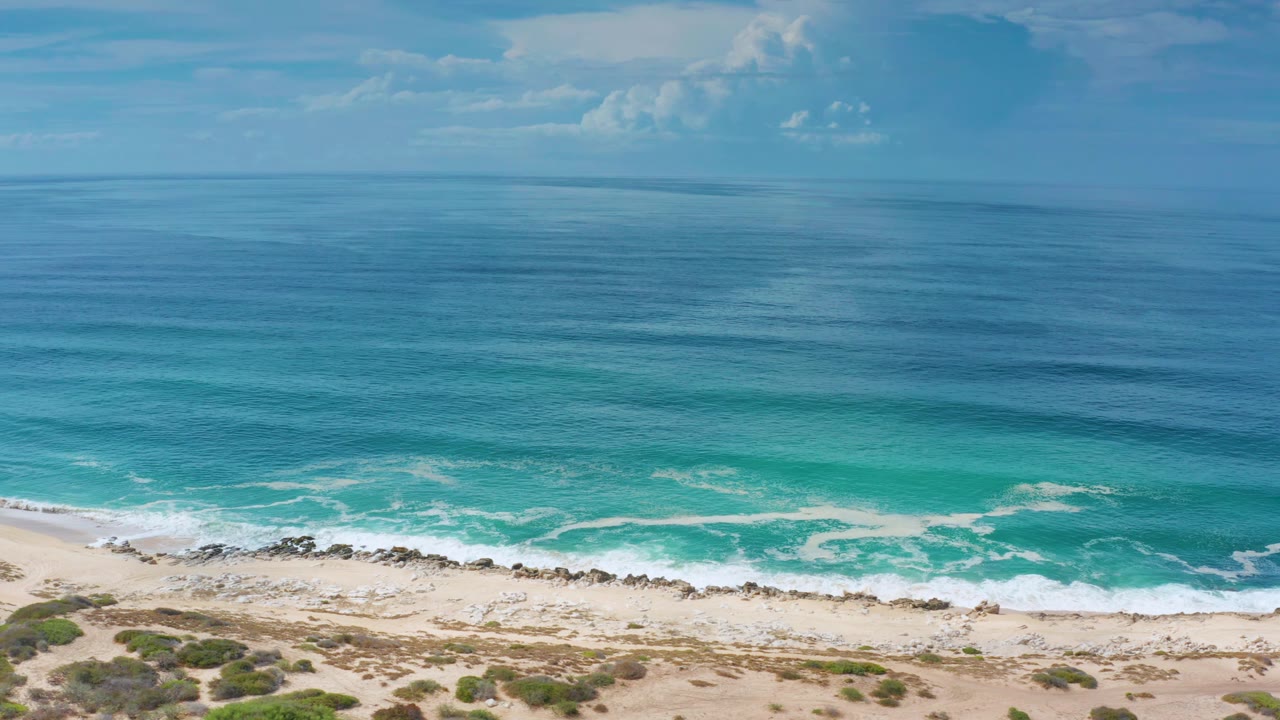 Open ocean, turquoise shallows, white surf, and a sandy beach under light clouds.