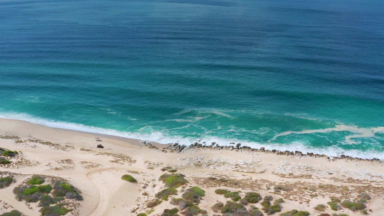 High view of the Los Cabos coast with deep blue ocean above turquoise surf, pale sand below, scattered rocks near the waterline, dune scrub in the foreground, and no visible people.