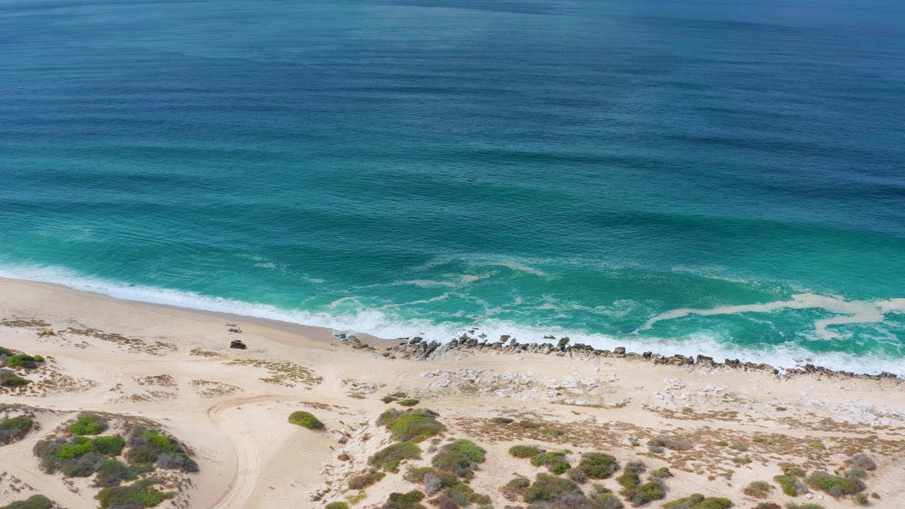 Los Cabos shoreline seen from above, with a broad band of blue ocean, white surf spreading over turquoise water, pale sand, a rocky strip, and no visible people.