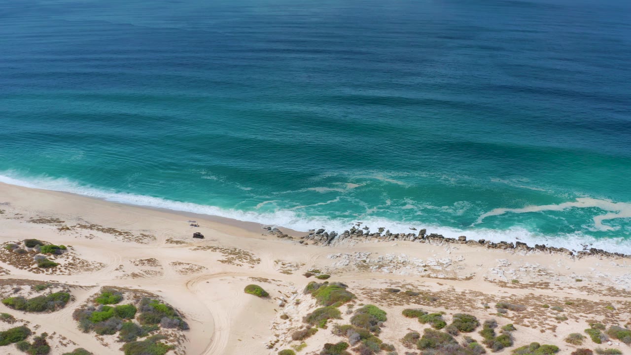 Aerial frame of the Los Cabos beach where white water reaches the sand beside a dark rocky line, with open ocean filling the upper frame and no visible people.