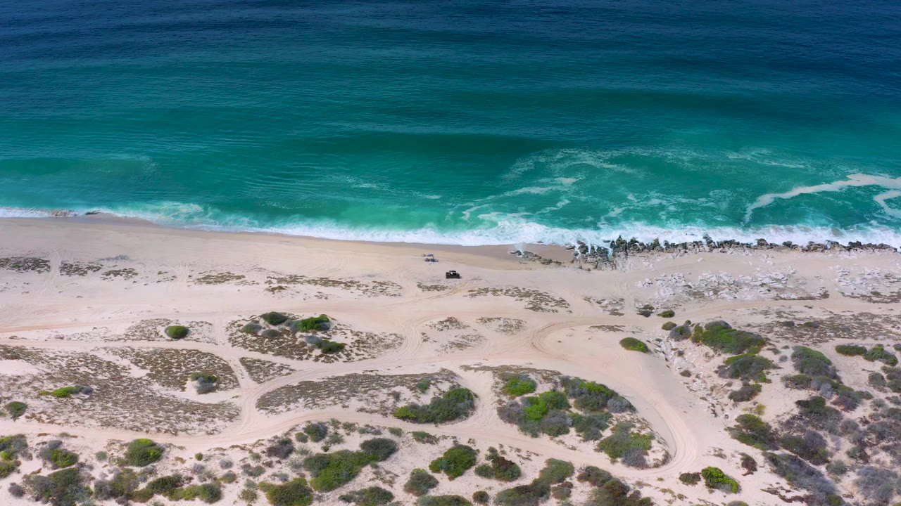 A Los Cabos aerial beach frame shows the surf line slightly farther along the sand, with white foam, scattered rocks near the water, low green shrubs, and no visible people.