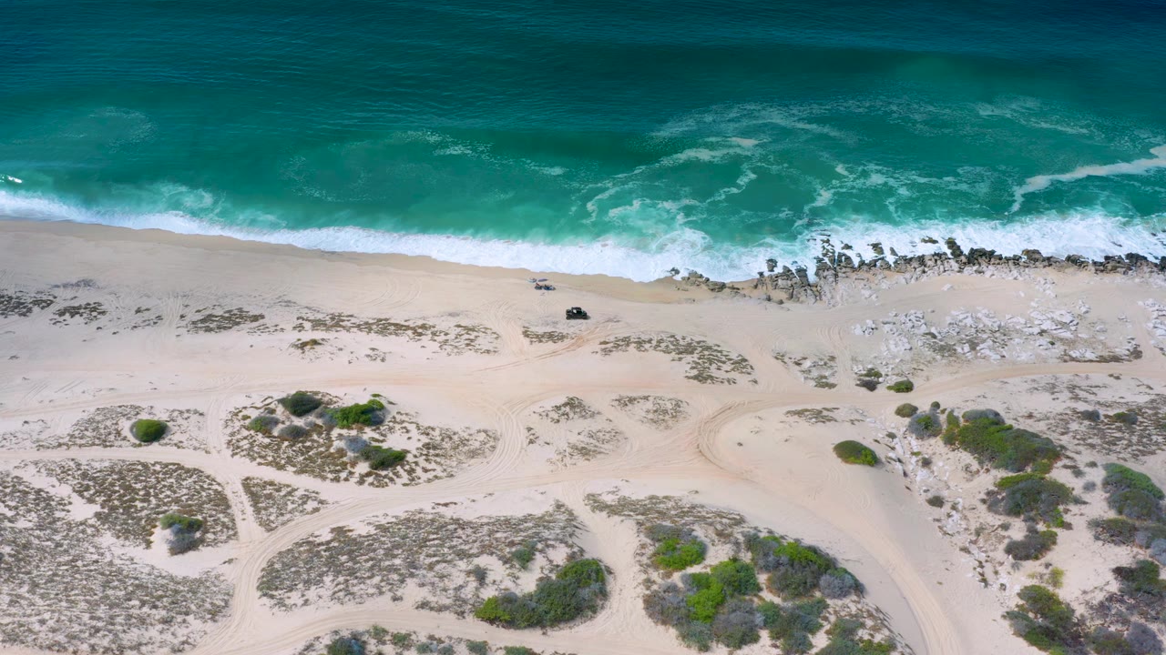 Aerial view over the Los Cabos shoreline with a band of breaking surf, darker rock patches at the waterline, pale sand, low shrubs, and no visible people.