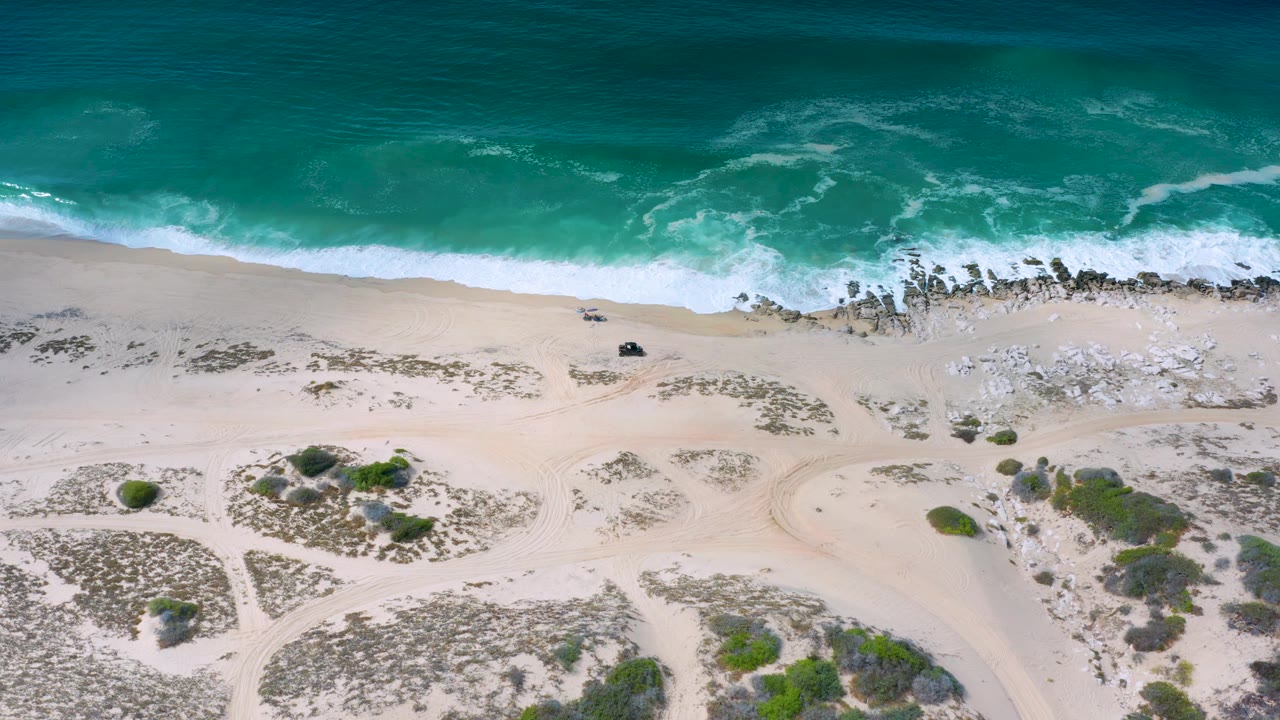 Top-down coastline with foamy surf, rocks, open sand, and scrub patches.