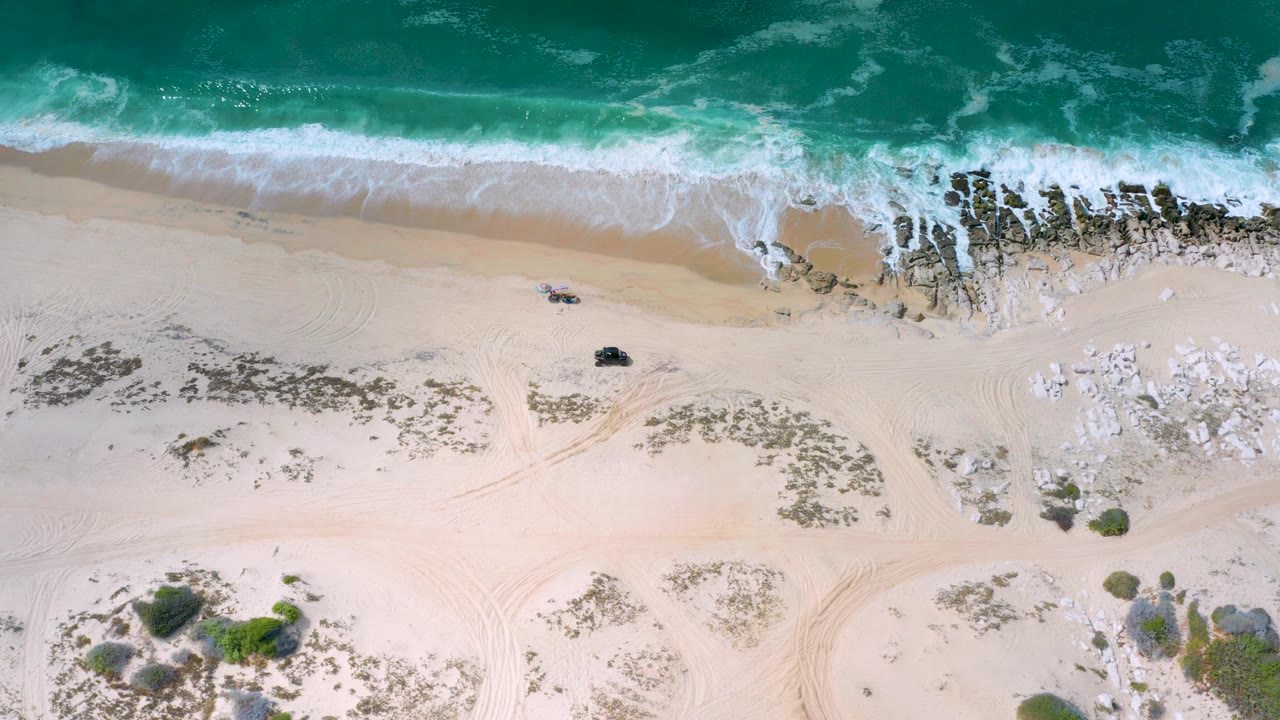 Overhead Los Cabos beach frame with turquoise surf at the top, pale sand below, dark shoreline rocks to the right, and tiny people visible near the waterline.