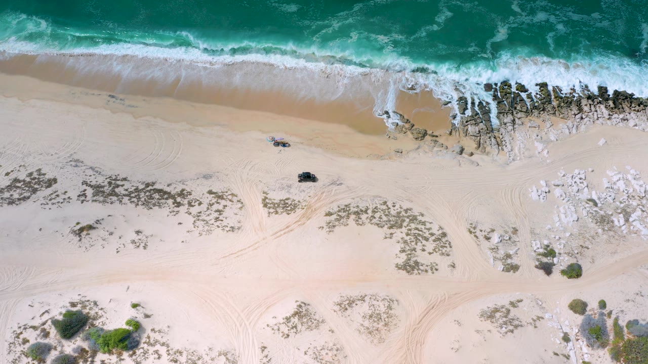 The Los Cabos coast appears from directly above with teal water, foamy surf, open pale sand, shoreline rocks, and tiny people near the upper sand area.