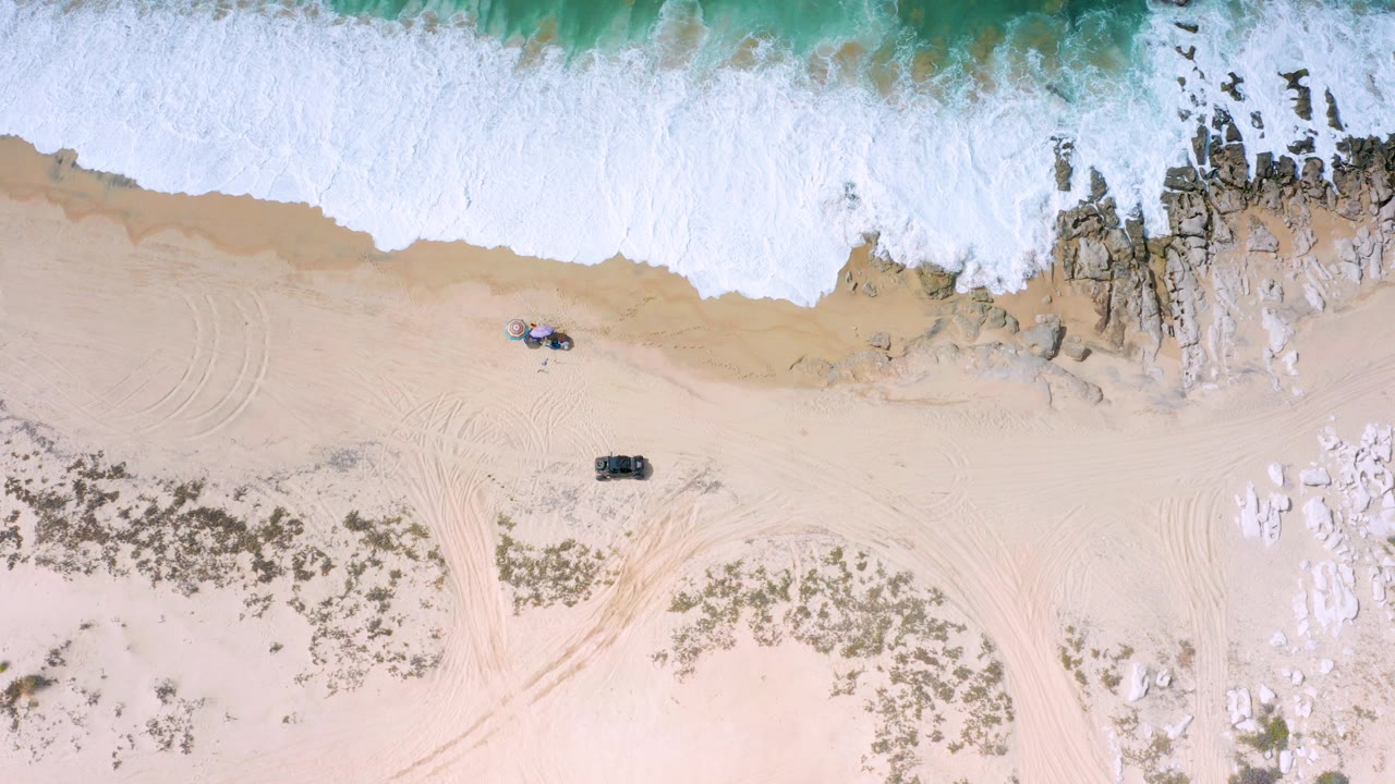 Overhead view of Los Cabos shoreline with white surf at the top edge, tan sand below, dark rocks near the waterline, faint tire tracks, and no visible people.