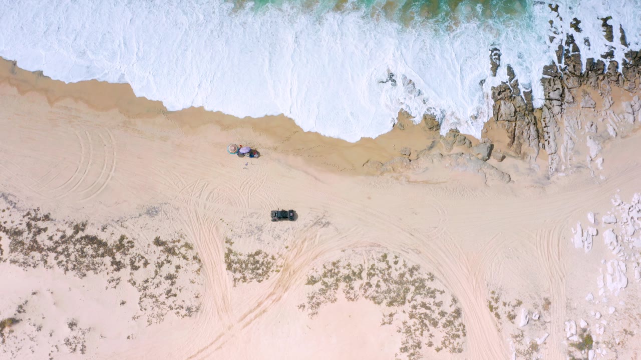 Overhead Los Cabos shoreline with broad white wash across the upper frame, dark rocks exposed near the surf, open sand below, curved track marks, and no visible people.
