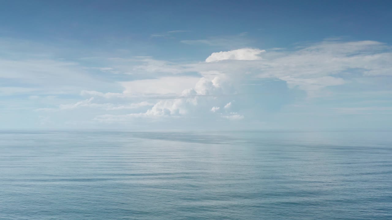 Mid-clip frame with the ocean stretching to a distant horizon and a larger white cloud formation centered in the Los Cabos sky, with no visible people.