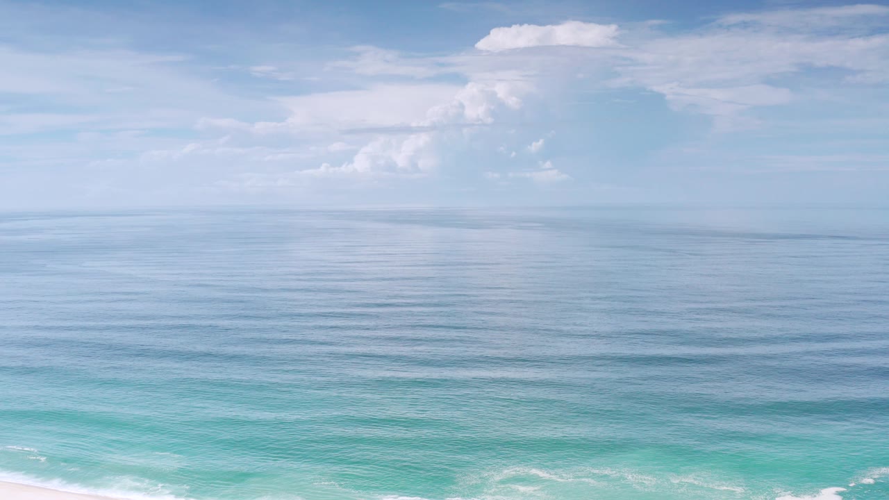 Opening frame of the Los Cabos, Mexico coast with pale sky, white clouds, calm blue water, turquoise shore break, a narrow sand edge, and no visible people.