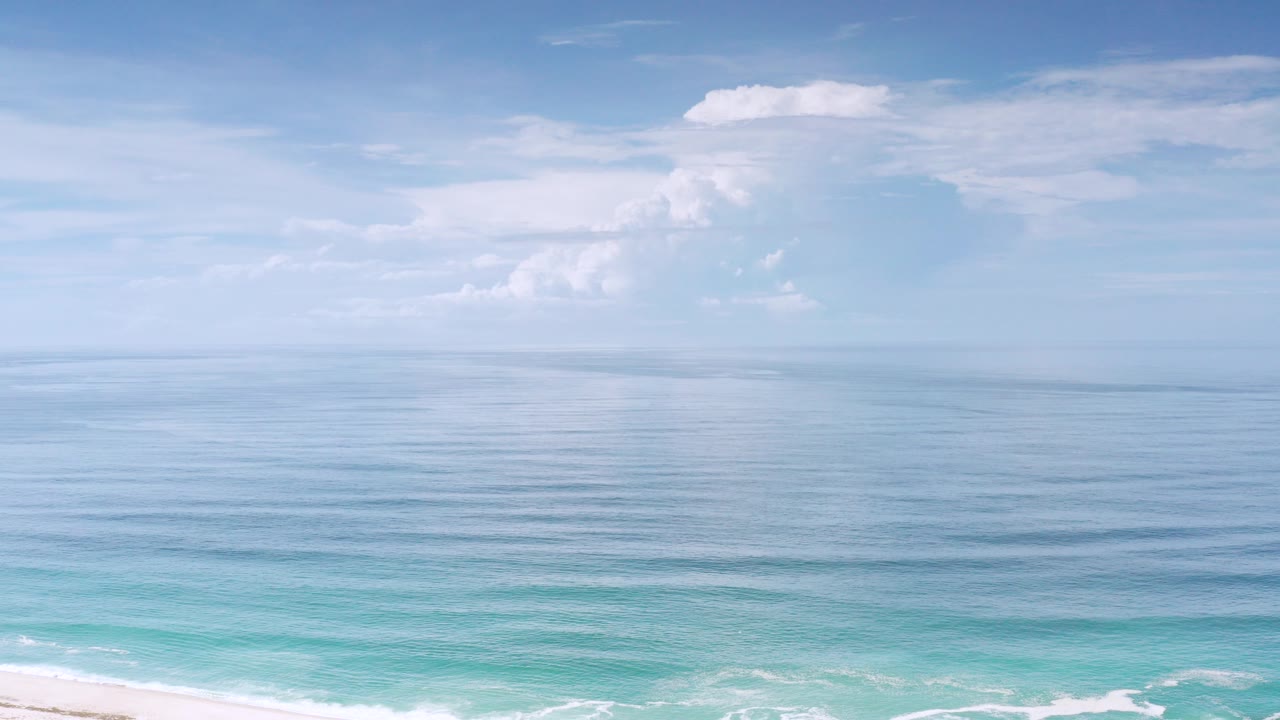 Opening frame of the Los Cabos clip showing calm blue ocean, a low horizon, pale sky, white clouds, turquoise shore water, and no visible people.
