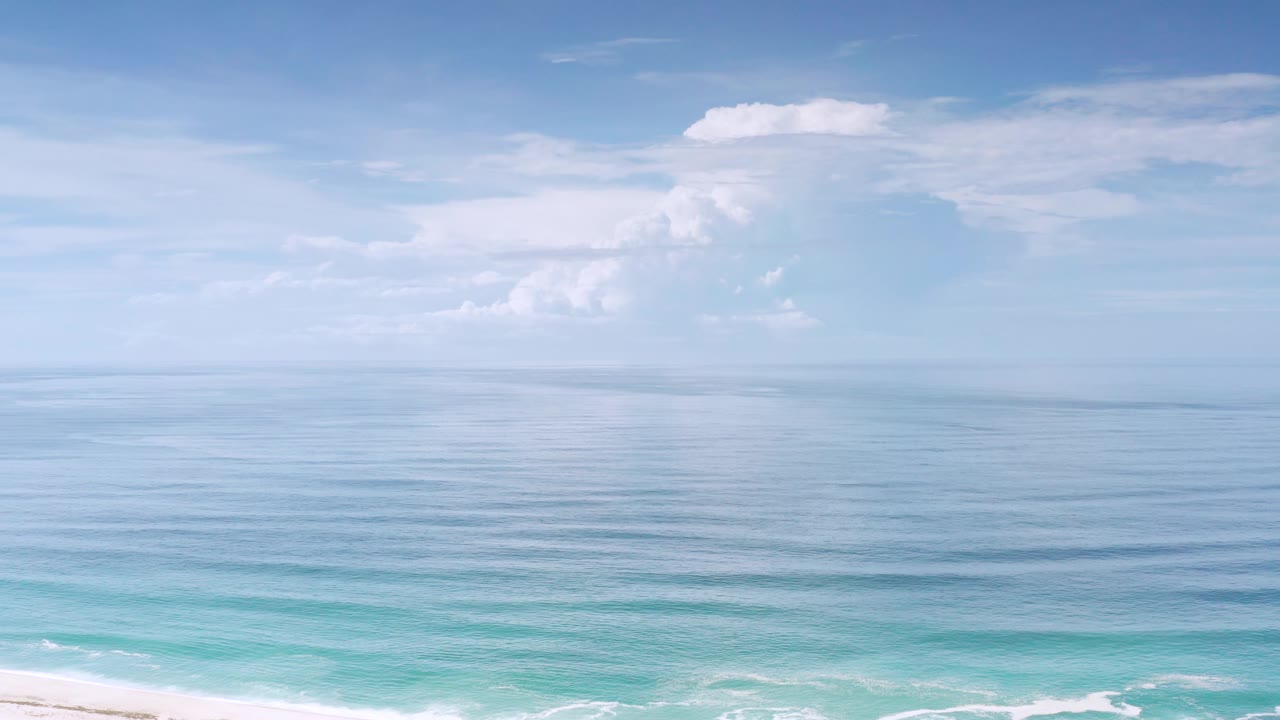 Mid-clip Los Cabos frame with smooth blue water below a wide bright sky, soft white clouds, a narrow surf line, and no visible people.