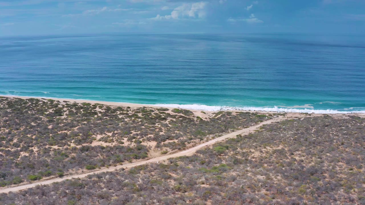Drone-height view in Los Cabos, Mexico showing turquoise nearshore water, thin white surf, open sand, dry coastal scrub, and no visible people.