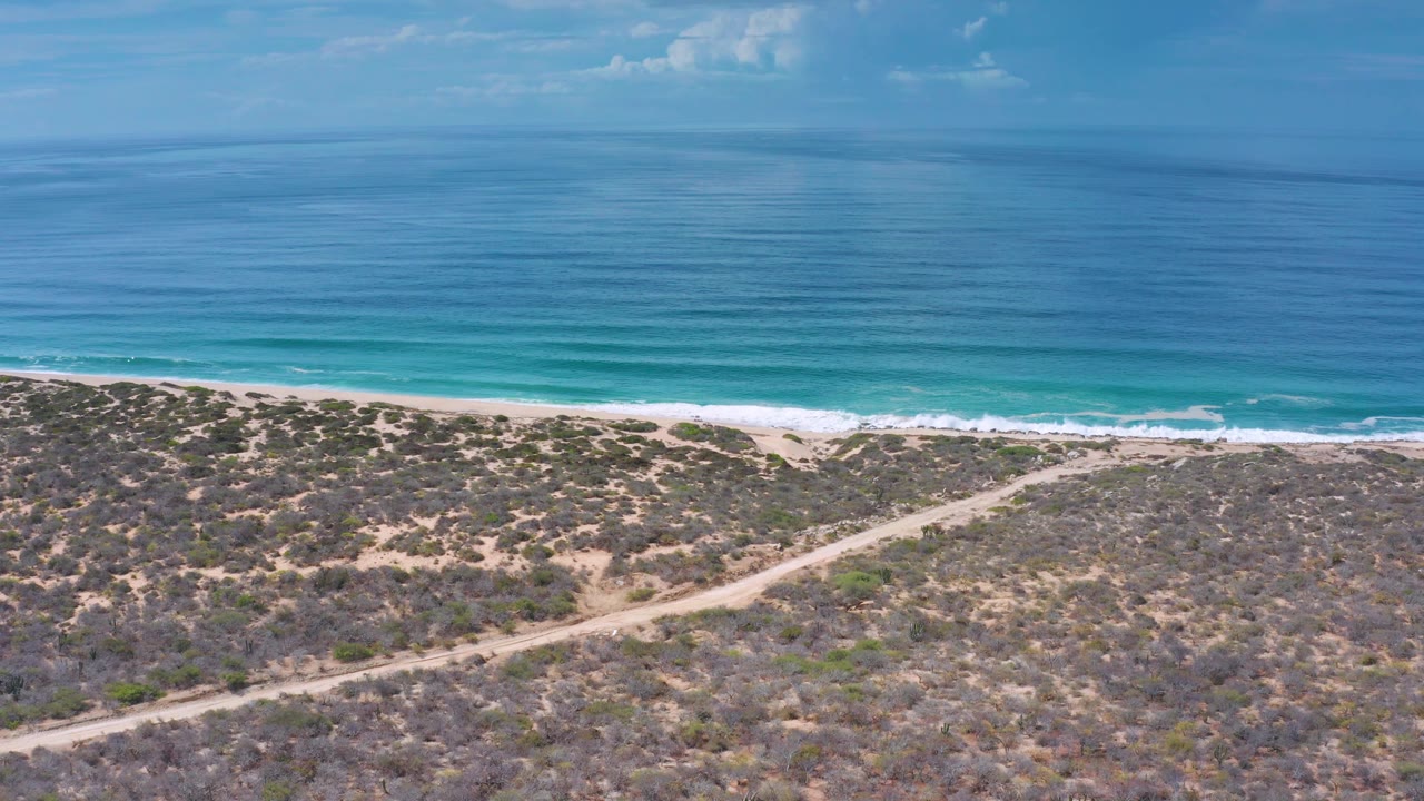 Wide aerial frame over Los Cabos, Mexico with a level ocean horizon, bright blue water, a narrow beach, scrubland, a diagonal sandy track, and no visible people.