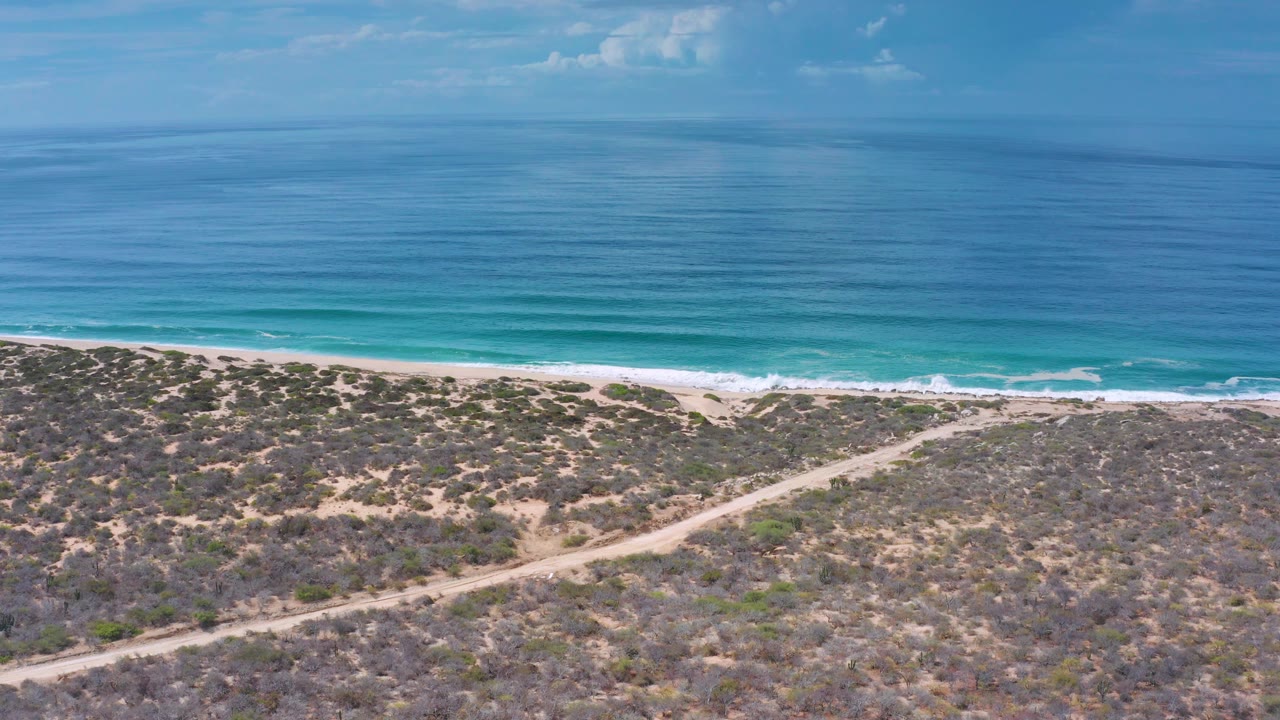 Aerial Los Cabos, Mexico view with calm blue ocean, white wave edges, a light sand shoreline, dry scrub terrain in front, and no visible people.