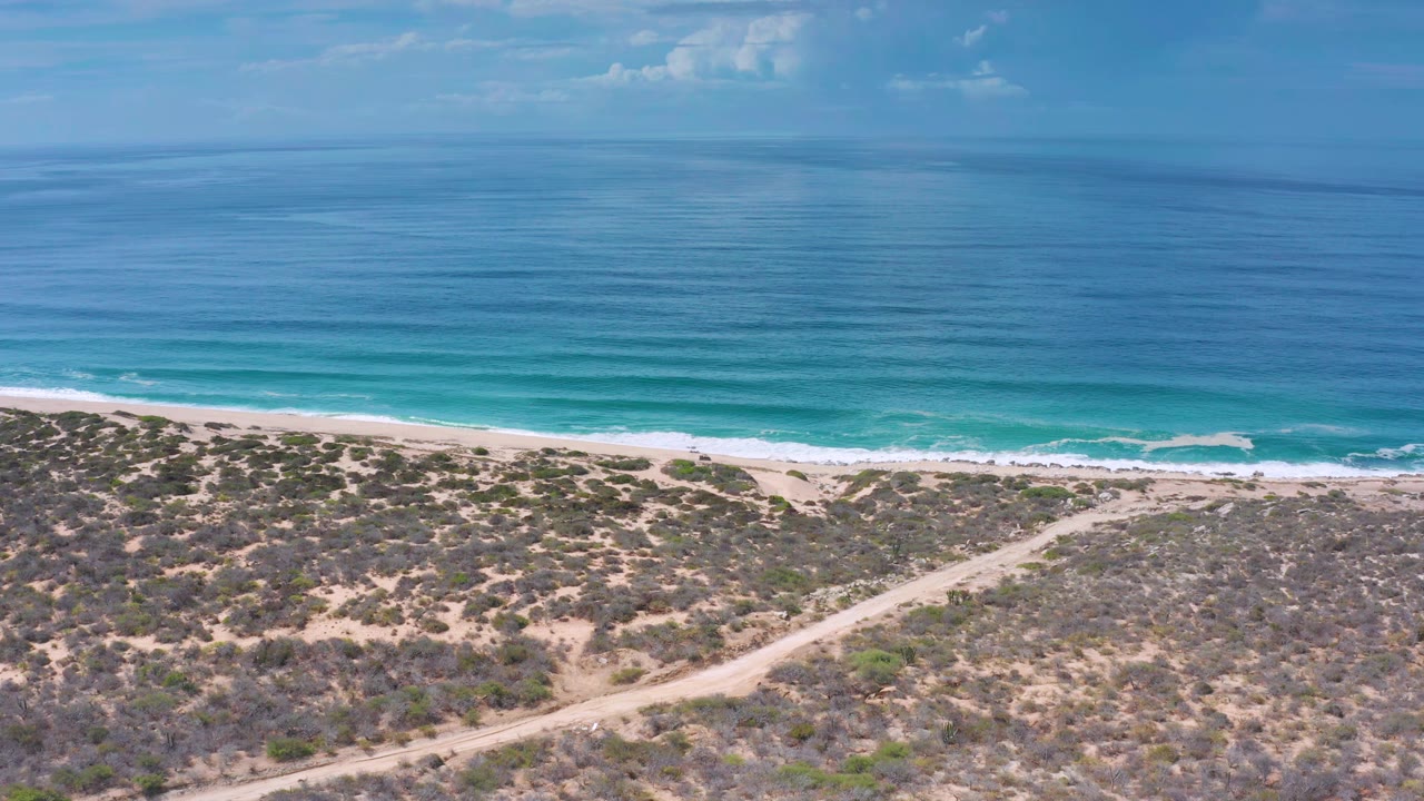Aerial frame over Los Cabos, Mexico, showing turquoise water bands, a narrow beach strip, low coastal scrub, a sandy road, and no visible people.