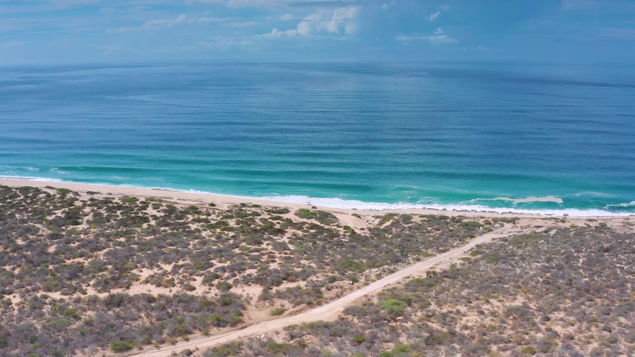 Aerial Los Cabos, Mexico view with calm blue sea, a thin surf break, scrub-covered dunes, a curving dirt track, and no visible people.