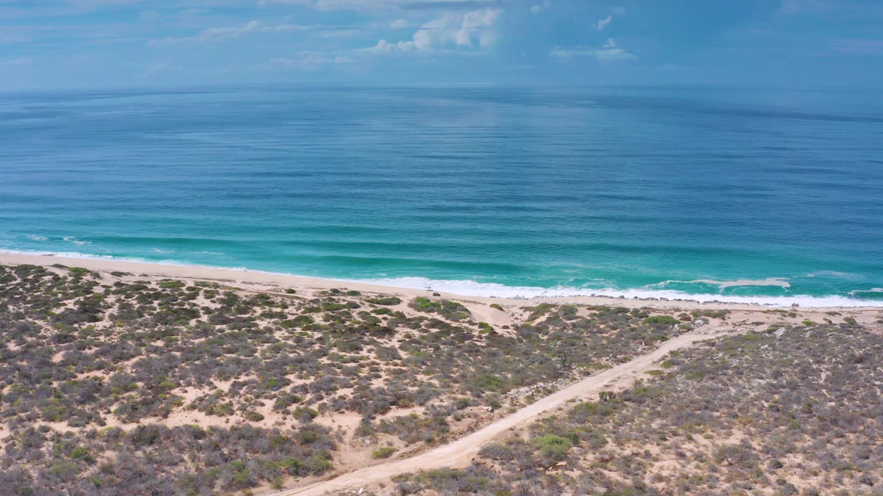 Mid-clip aerial frame from Los Cabos, Mexico, with broad blue water, white waves along the beach, patchy scrub in the foreground, pale sand, and no visible people.