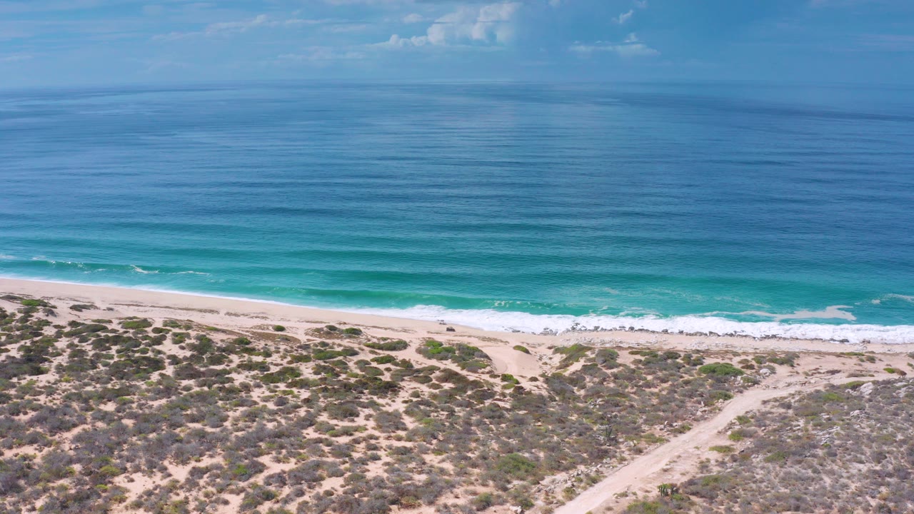 Opening aerial frame over Los Cabos shoreline with blue ocean bands, a thin surf line, pale beach, scrub dunes, and no visible people.
