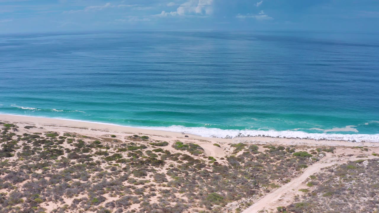 Los Cabos aerial frame showing calm blue water, a narrow surf break, pale sand, scrub patches, a diagonal sand path, and no visible people.