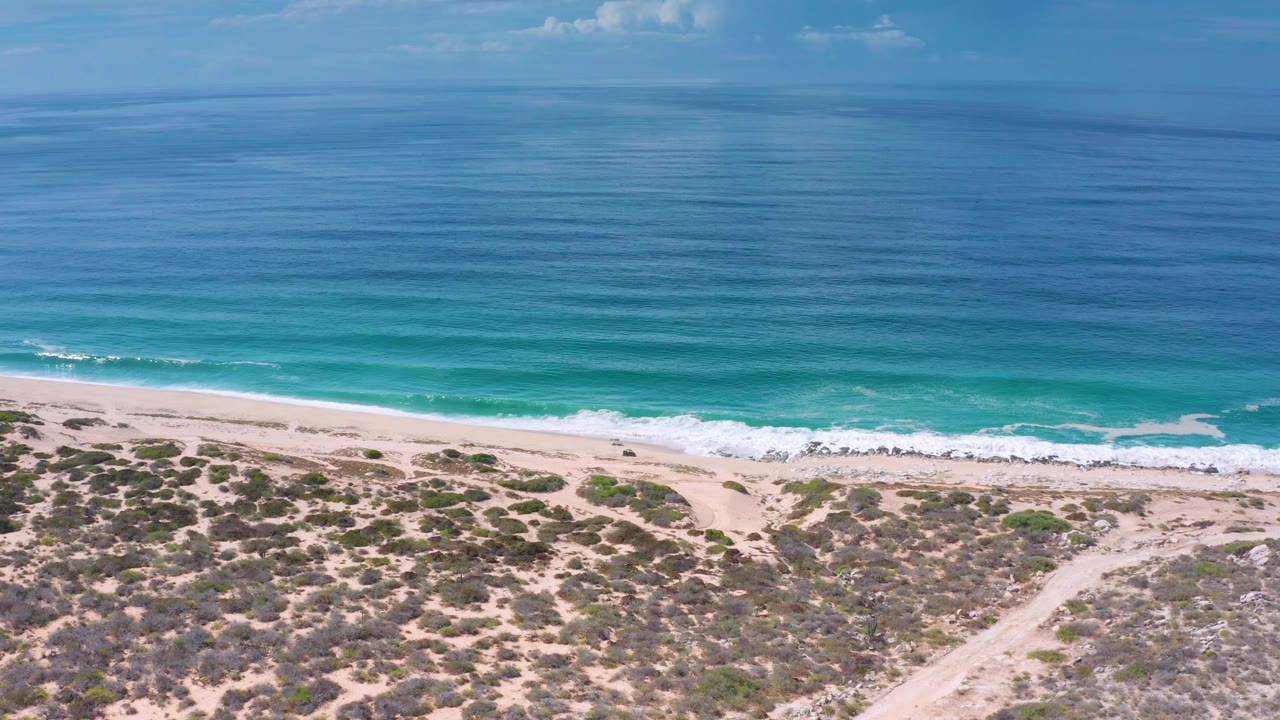 Mid-clip aerial frame over Los Cabos with deep blue water fading to turquoise shallows, a foamy shore break, scrub-lined sand, and no visible people.