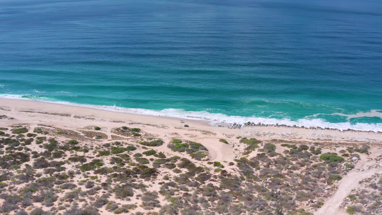 Los Cabos aerial view showing a broad band of ocean, a thin line of breaking waves, light sand, scrub patches, and no visible people.