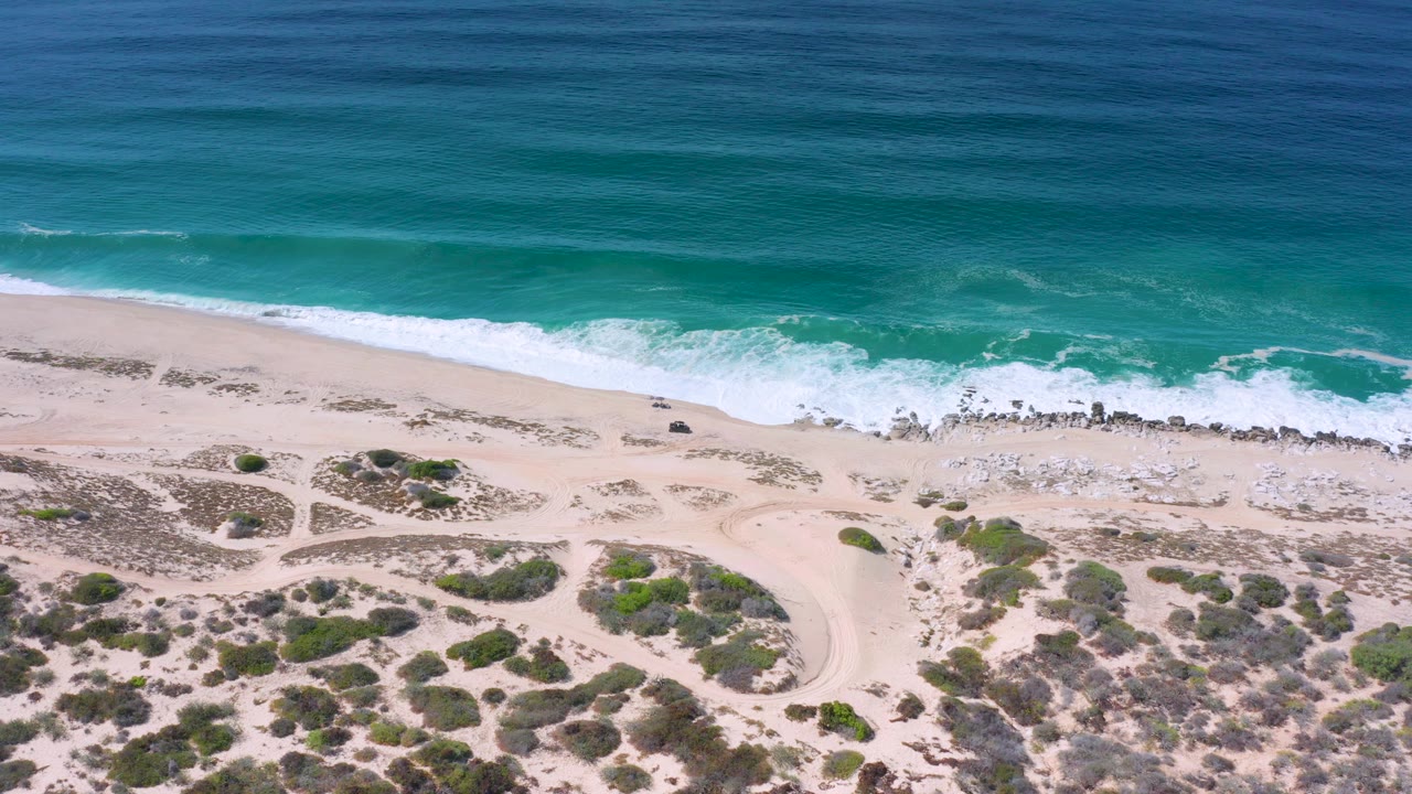 Overhead coastline with green-blue water, white surf, sand, rocks, and dune scrub.