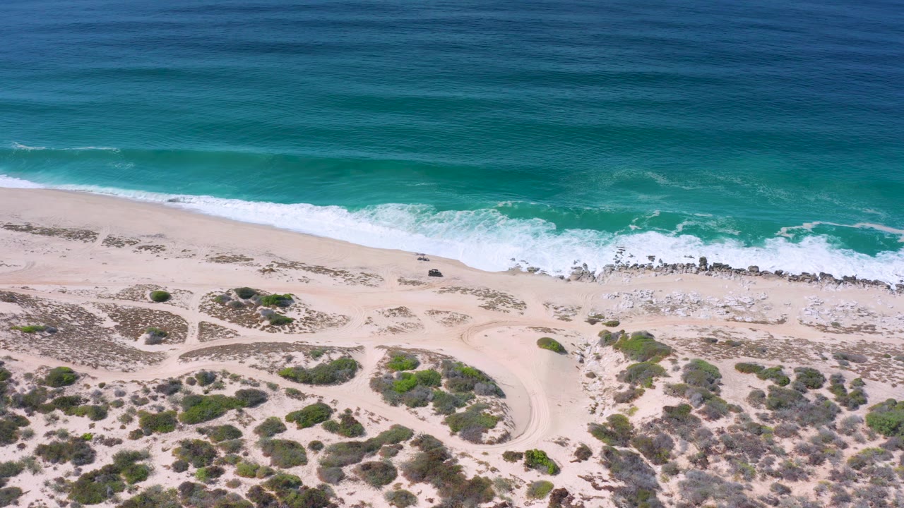 Los Cabos aerial frame with turquoise water, a white wave band, exposed rocks near the shore, branching sandy tracks, scattered scrub, and no visible people.