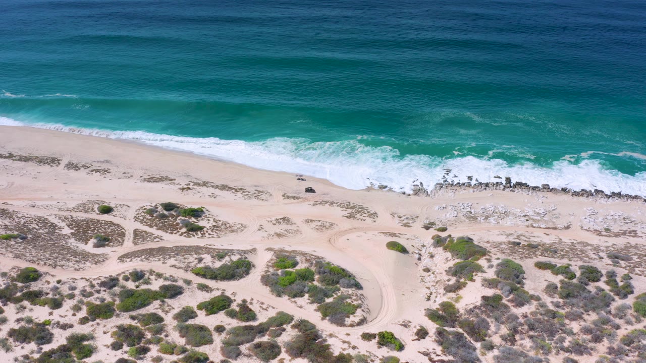 Top-down coastal view in Los Cabos with darker blue water farther out, green shallows, foamy surf over rocks, pale sand, dune vegetation, and no visible people.