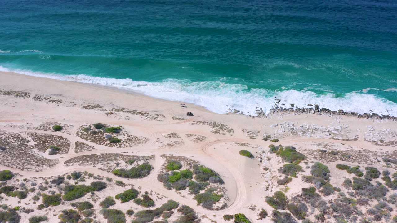 Wide overhead frame from Los Cabos showing a long band of surf crossing a rocky shoreline, pale open sand, scrub patches, sandy paths, and no visible people.